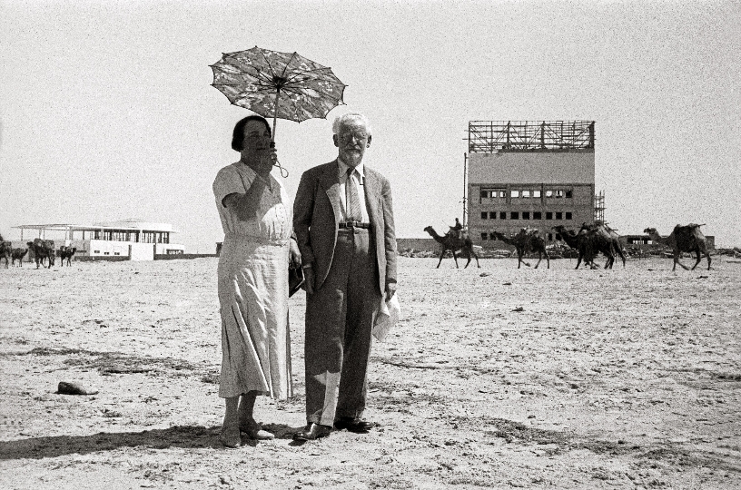 Paar steht vor Wüste und hält einen Schirm, im Hintegrund laufen Kamele vor Gebäuden