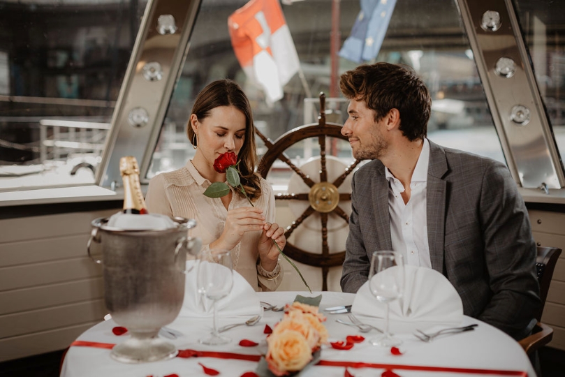 Frau riecht im Beisein ihres Begleiters an roter Rose im romantischen Dinner-Ambiente eines Flusskreuzfahrtschiffes